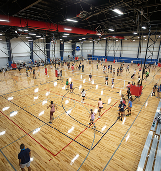 A large indoor gym with several basketball courts, filled with people playing and watching games. Bright lighting and a lively, energetic atmosphere.