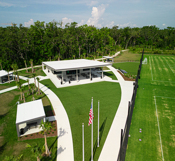 Aerial view of a sports complex showing a modern pavilion with seating on a manicured lawn. Nearby are flagpoles and a soccer field, bordered by lush trees.