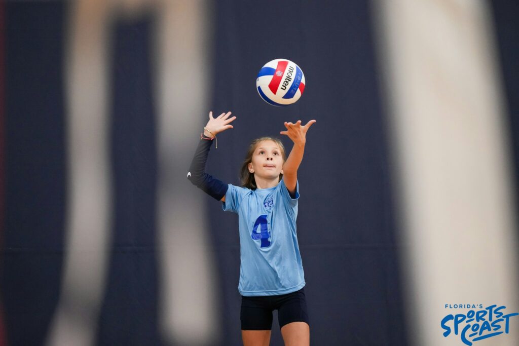 A girl in a blue shirt and black shorts strikes a volleyball during a game on a sandy court
