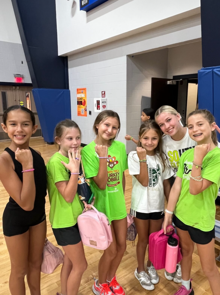 A group of girls wearing green shirts and pink backpacks smile together for a photo.