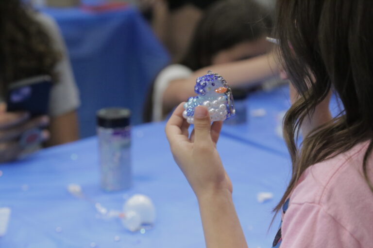 A girl sits at a table, holding a small plastic bird in her hand, smiling at the camera.