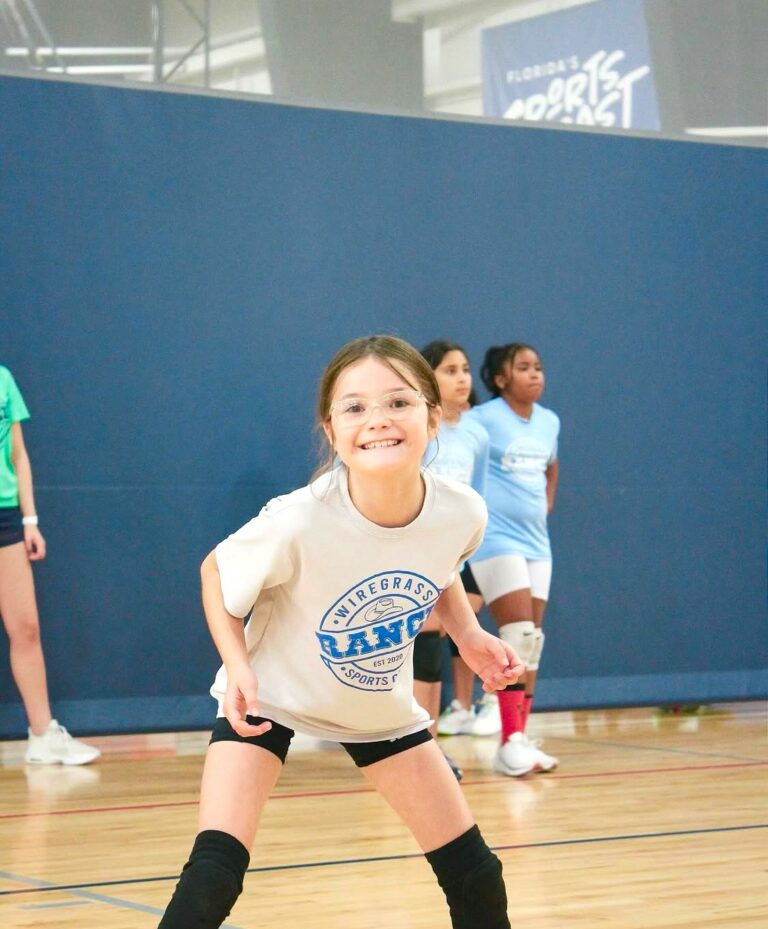 A young girl in a blue shirt holds a volleyball, smiling and ready to play.