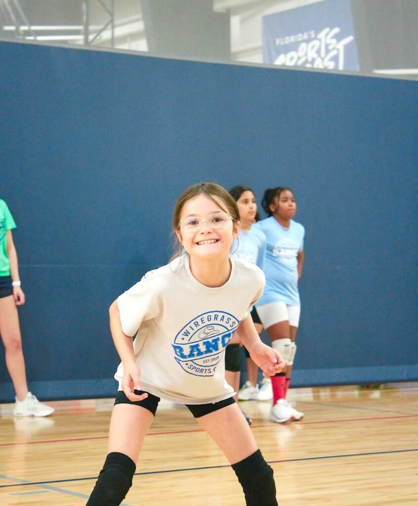 A young girl in a blue shirt holds a volleyball, smiling and ready to play.