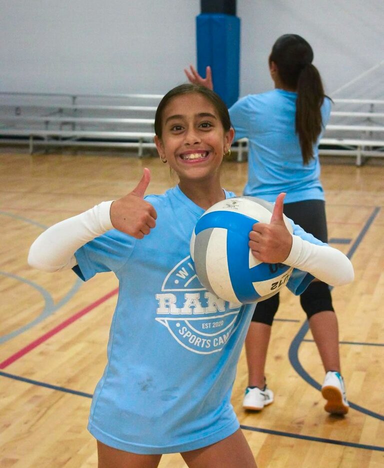 A young girl in a blue shirt holds a volleyball, smiling and ready to play.