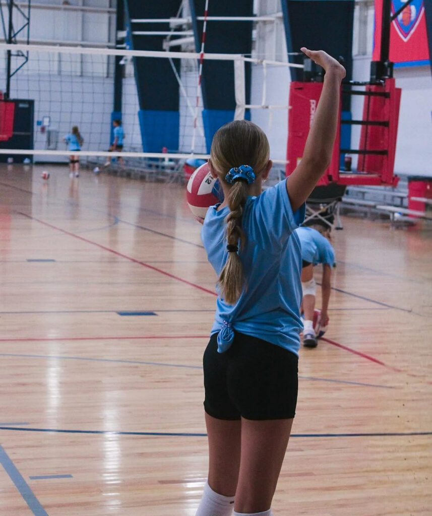 A young girl in a blue shirt holds a volleyball, ready to play.