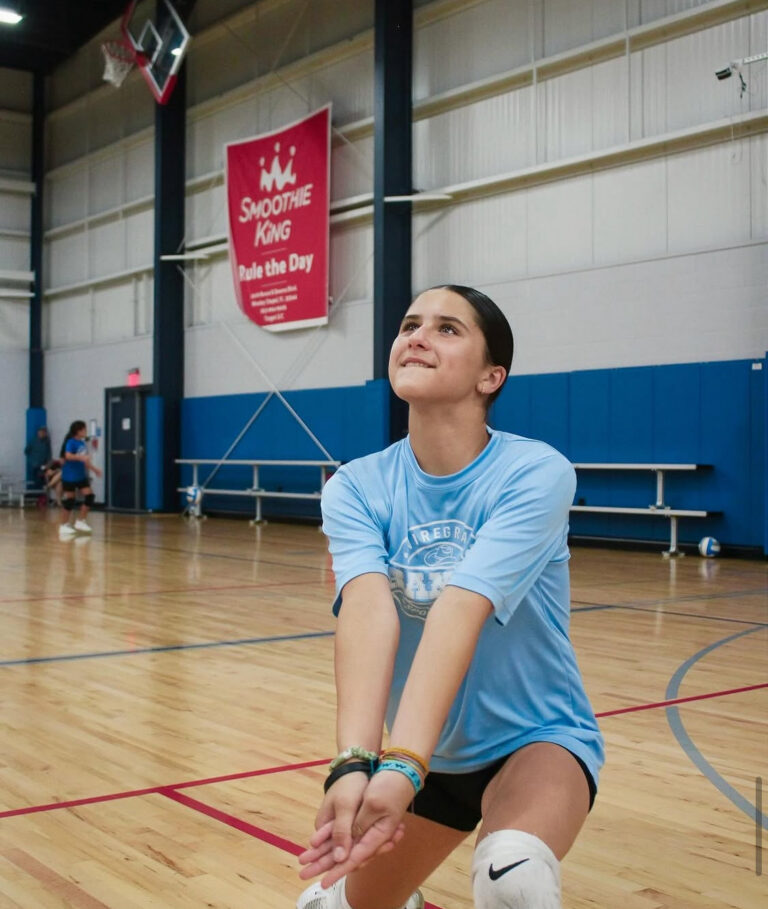 A girl in a blue shirt and black shorts strikes a volleyball during a game on a sandy court