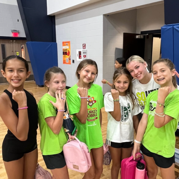 A group of girls wearing green shirts and pink backpacks smile together for a photo.