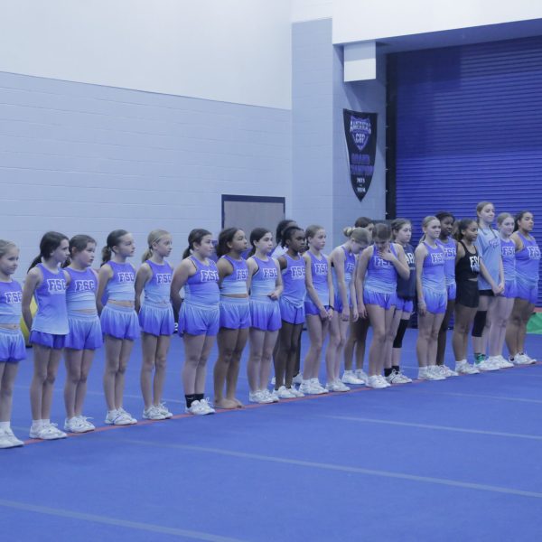 A cheerleading team stands in a line, wearing uniforms, inside a gym with banners and mats in the background.